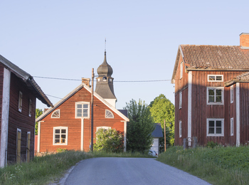 Landsväg mellan röda trähus och en kyrkobyggnad med torn i en lantlig miljö.