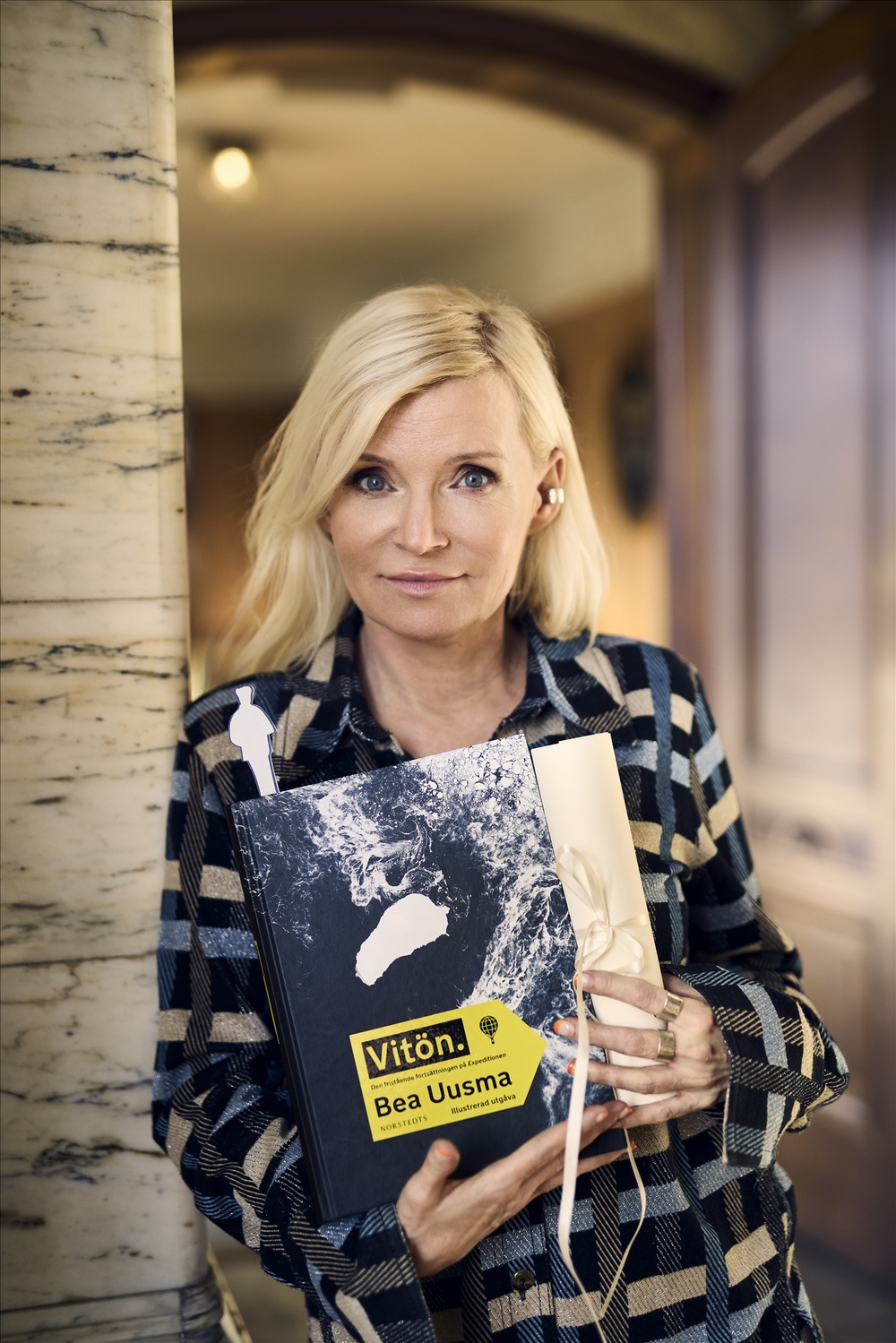 Person med blont hår håller boken 'Vitön' av Bea Uusma och ett inslaget paket i en hall.