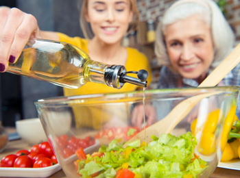 Två kvinnor lagar mat tillsammans och häller olja i en salladsskål med grönsaker.