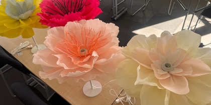 Fyra stora, färgglada konstgjorda blommor på lampfötter står på ett bord i ett mötesrum.