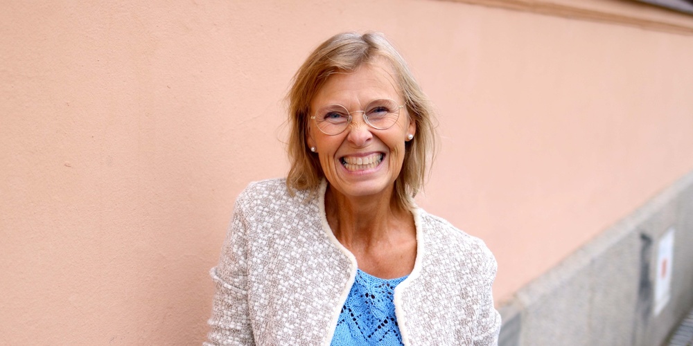 Leende äldre kvinna med glasögon står mot en ljusrosa vägg utomhus.