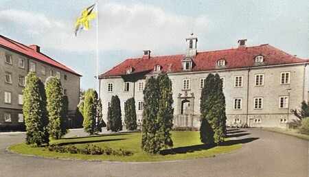 Stort stenhus med rött tak, flaggstång och rund plantering med höga träd framför byggnaden.