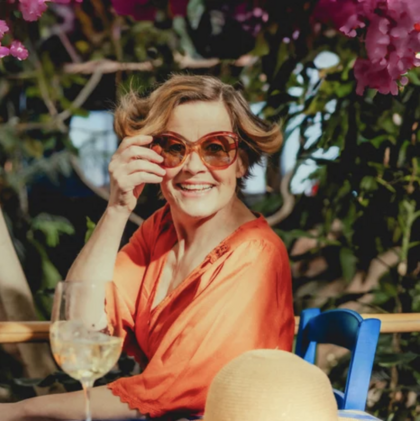 Kvinna i orange klänning sitter utomhus med stora solglasögon, leende, omgiven av blommor och ett glas vin.