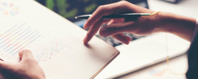 En person granskar diagram och grafer på ett papper med en penna, med en bärbar dator i bakgrunden.