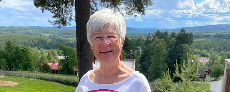 Äldre kvinna med vitt hår och glasögon står utomhus i vit klänning med röda detaljer, leende mot kameran.