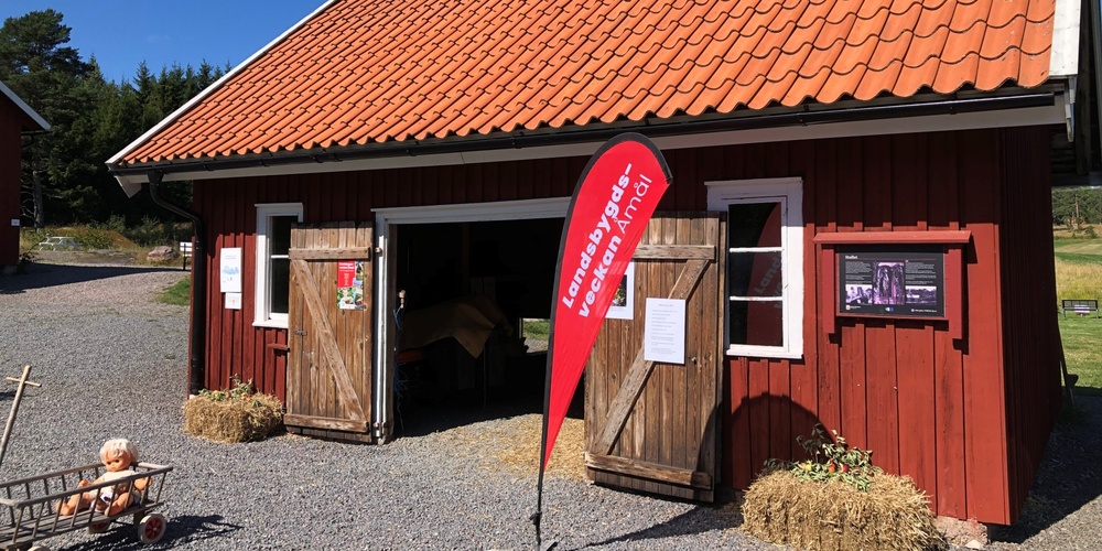 Röd träbyggnad med öppna dörrar, höbalar, skyltar och en docka i en vagn utomhus en solig dag.