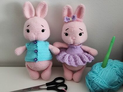 Två virkade kaniner i pastellfärger, en med blå väst och en med lila klänning, står bredvid garn och sax.