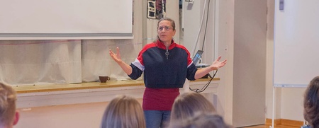En kvinna står framför en grupp och håller en presentation i ett klassrum med projektor och whiteboard.