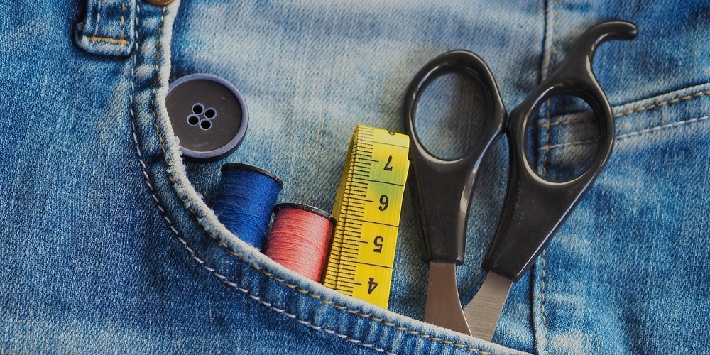 Knapp, måttband, tråd och sax instucket i en bakficka på ett par jeans.