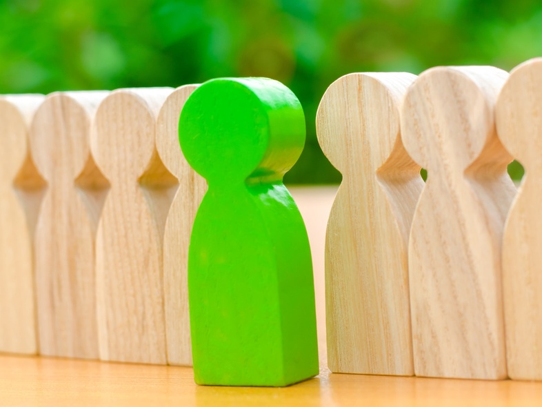 En grön träfigur står mellan två rader av ljusa träfigurer på ett bord med grön bakgrund.