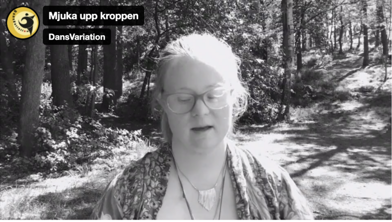 Person med långt hår står i en solig skogsmiljö, iförd mönstrad sjal och halsband.