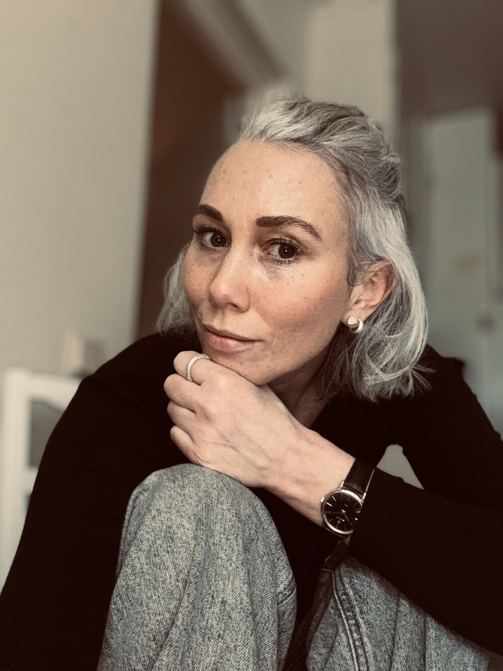 Person med grått hår sitter i ett rum, ansiktet är suddat för anonymitet. Bär svart tröja, grå byxor och klocka.