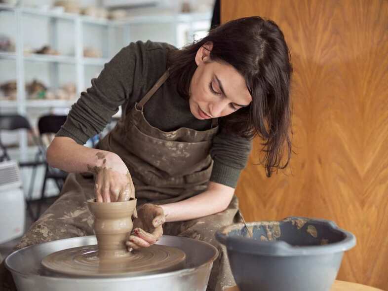 En person sitter vid en drejskiva och arbetar på en vas i lera.