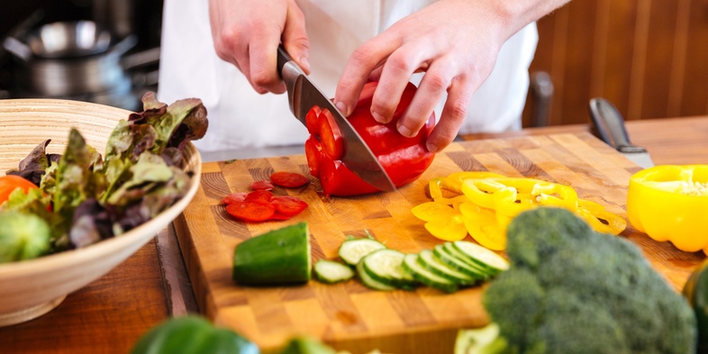 En person skär röda paprikor på en skärbräda omgiven av olika grönsaker i ett kök.