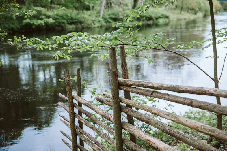 En trästaket står vid kanten av en lugn flod omgiven av grönskande natur.