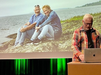 Person i rutig skjorta står vid ett podium med en dator som visar en stor bild på projektorduk i bakgrunden. På den bilden är det två personer som sitter på en sten vid vatten.