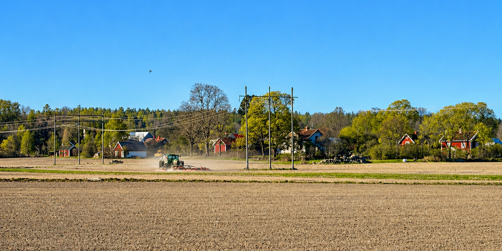 En traktor arbetar på en åker med bondgårdar och gröna träd i bakgrunden under en klarblå himmel.