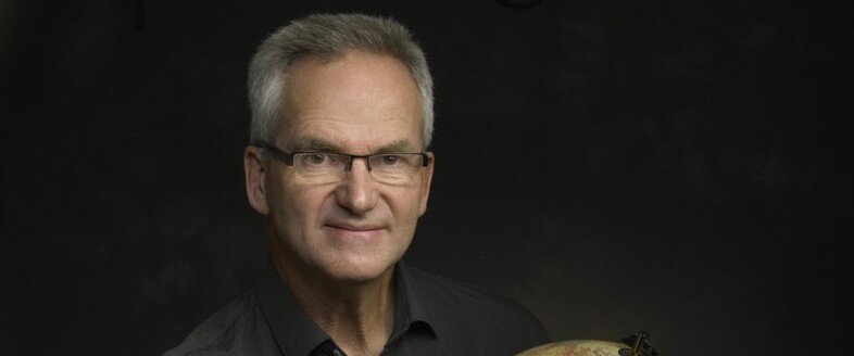 Äldre man med glasögon och grått hår poserar mot mörk bakgrund, håller en jordglob.