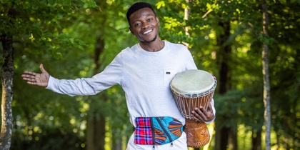 En leende man står barfota på en trästig i skogen och håller en trumma under armen.