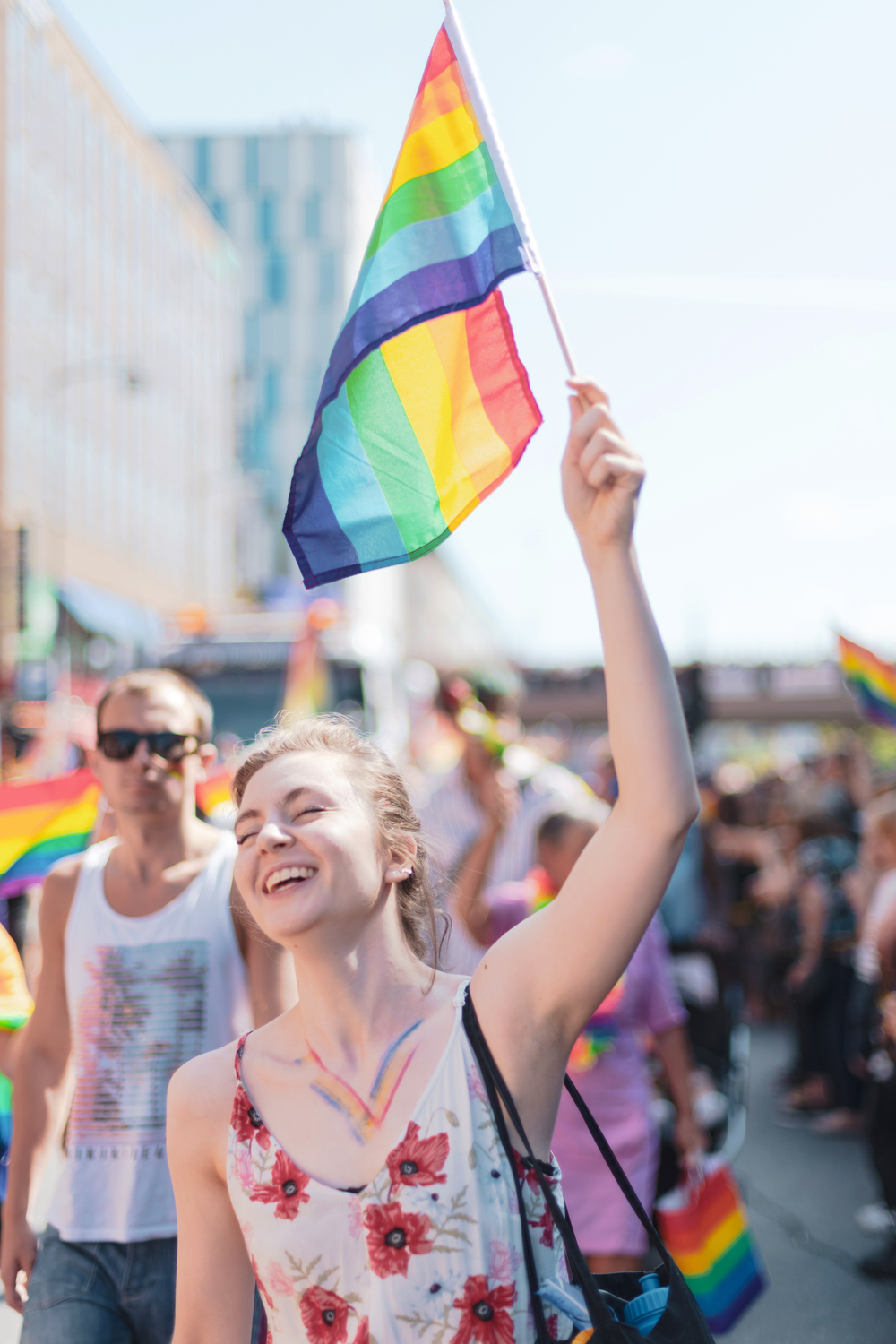 Leende person håller en regnbågsflagga på en prideparad under en solig dag.