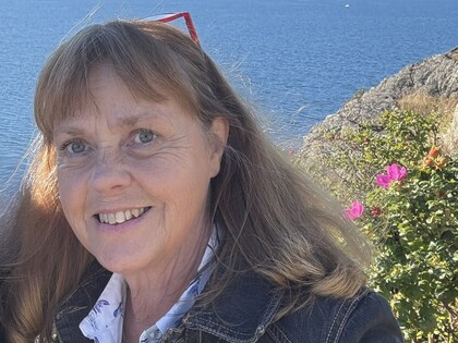 Person med långt hår står utomhus vid vatten och klippor, blommor syns i bakgrunden.