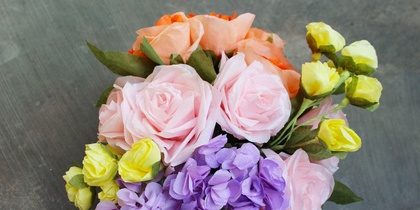 En bukett med färgglada konstgjorda blommor i rosa, lila, gult och orange på en grå bakgrund.