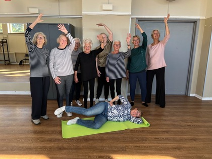 En grupp äldre kvinnor poserar glatt i ett ljust rum, en kvinna ligger på en grön yogamatta på golvet.