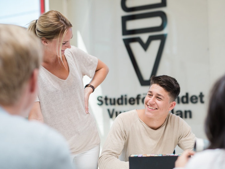 Fyra unga vuxna sitter och pratar runt ett bord i ett ljust rum med stora fönster.