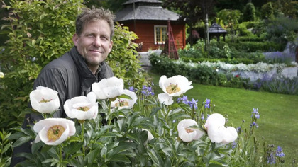Man står i en trädgård bredvid vita blommor, med en röd stuga i bakgrunden.