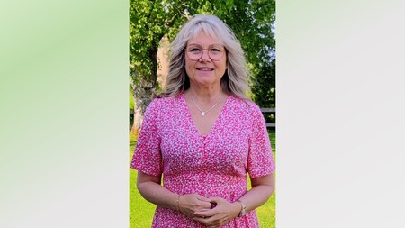 Kvinna med blont hår i rosa blommig klänning står utomhus på gräsmatta med träd i bakgrunden.