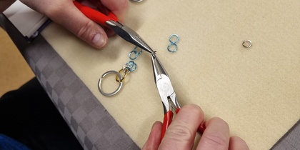 En person använder två tänger för att arbeta med små metallringar och skapa smycken på ett bord.