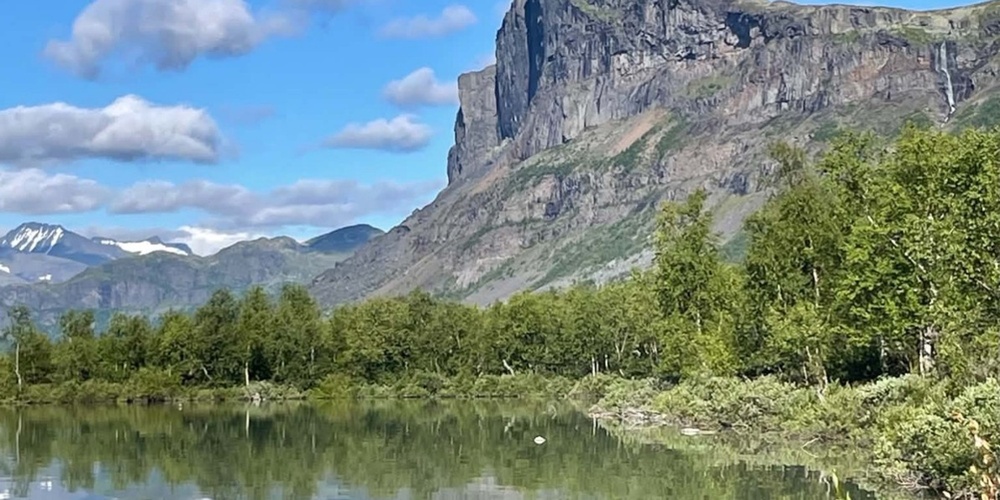Storslagen bergstopp speglas i en lugn sjö, omgiven av grönska och blå himmel med moln.