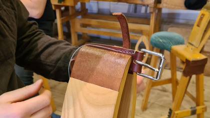 En person arbetar med att sy fast ett brunt läderbälte på en träbit i en verkstadsmiljö.