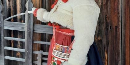 Kvinna i traditionell folkdräkt står vid en trägrind framför en gammal trävägg.