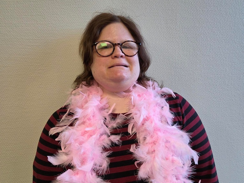 Person med randig tröja och rosa fjäderboa står mot en ljus vägg. Ansiktet är suddat.