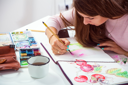 Kvinna målar akvarell i en skissbok vid ett bord med färger och en kopp te.