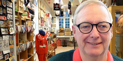 Äldre man med glasögon ler i ett rum fullt av samlarföremål och memorabilia.