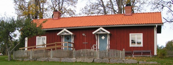 Rött trähus med vita knutar, två blå dörrar, staket och tegeltak, omgiven av träd och gräsmatta.