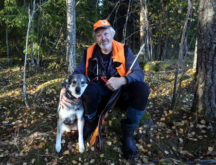 En man i jaktkläder sitter i skogen med en hund och ett gevär, omgiven av höstlöv.