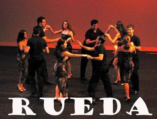 En grupp människor dansar i en cirkel på en scen med texten 'RUEDA' längst ner.