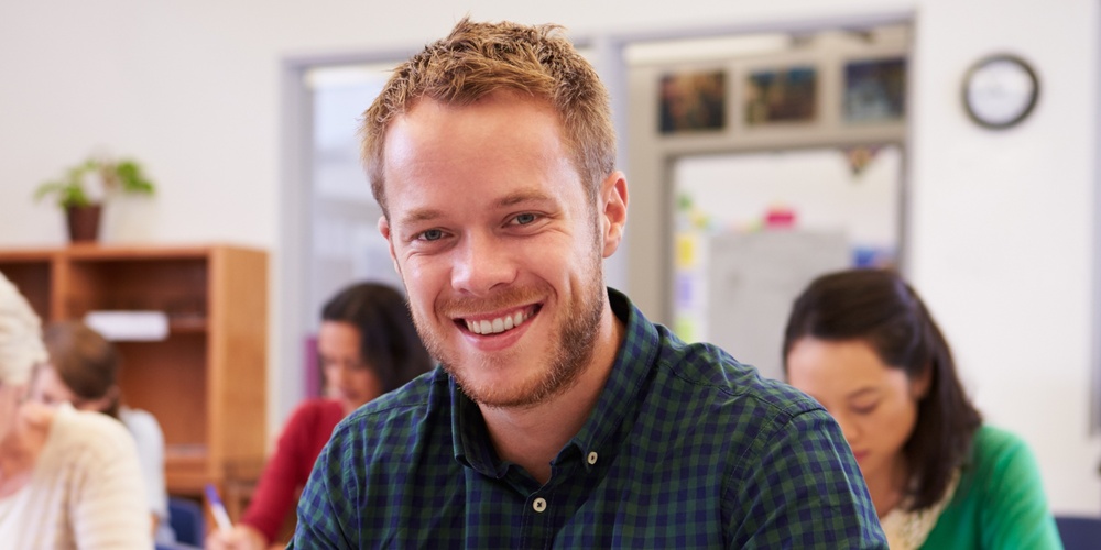 Leende man sitter vid skolbänk i klassrum, skriver i anteckningsbok. Andra vuxna studenter i bakgrunden.