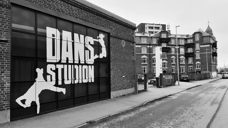 Svartvit bild på en dansstudio med stora fönster och texten 'DANS STUDIO' samt siluetter av dansare. Gata och äldre byggnader syns.