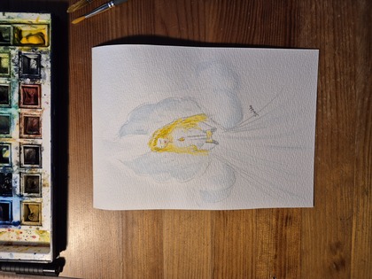 Akvarellmålning av en ängel med långt gult hår och vita vingar, placerad på ett träbord med färglåda och penslar.