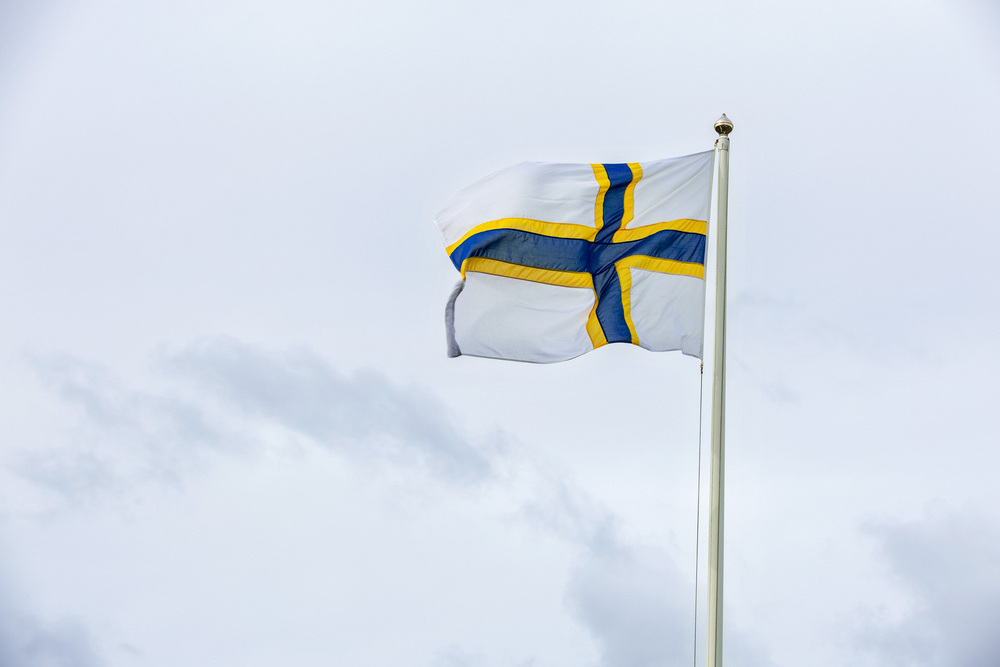 Flagga med blått kors och gul kant på vit bakgrund vajar mot en molnig himmel.