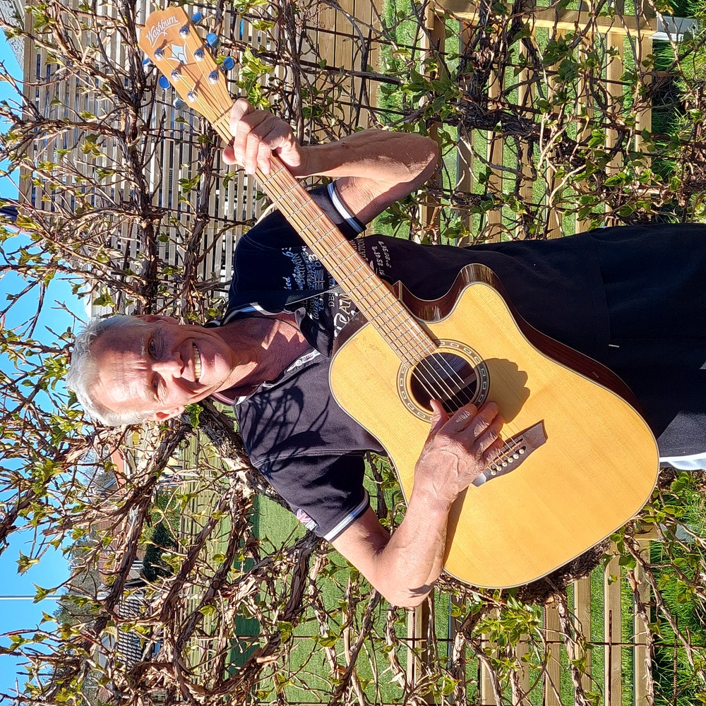 En äldre man står utomhus och spelar akustisk gitarr framför en grönskande buske.