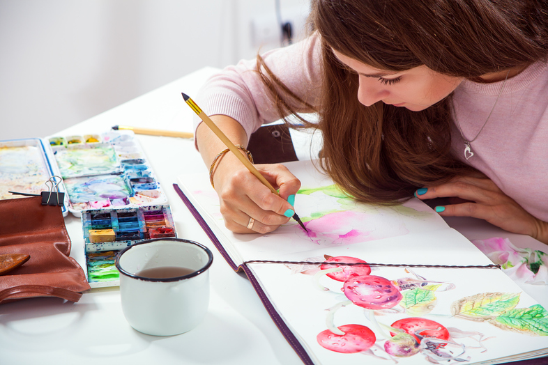 Kvinna målar akvarell i en skissbok vid ett bord med färger och en kopp te.