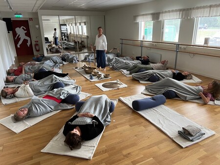 En grupp människor ligger på yogamattor med filtar i en ljus sal, en instruktör står i mitten.