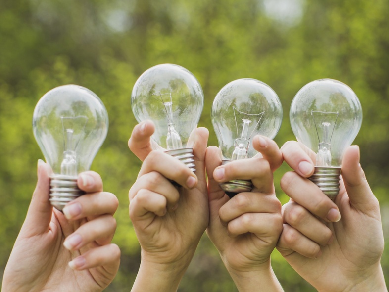 Fyra händer håller upp glödlampor utomhus med grönskande bakgrund.