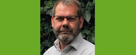 Man med glasögon och skägg står framför en bakgrund av gröna blad, bär skjorta med rutmönster.