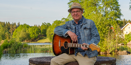 En person sitter utomhus på en rund bänk och spelar akustisk gitarr vid en sjö.